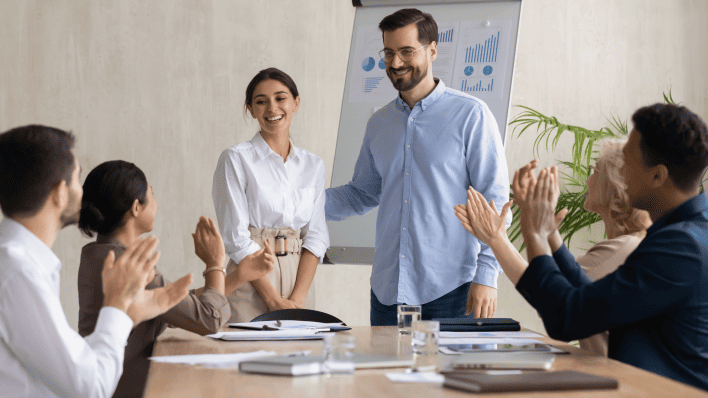 White bearded man in blue dress shirt is introducing a dark haired woman in a white blouse in a conference room. She is smiling and others are clapping.