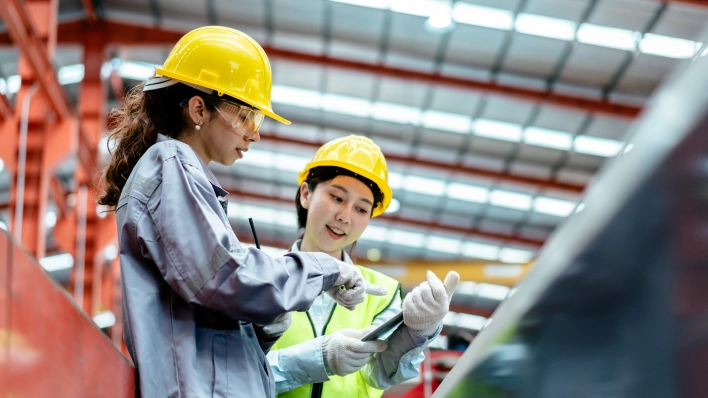 Two female production workers wearing hard hats and gloves pointing at a tablet screen. 