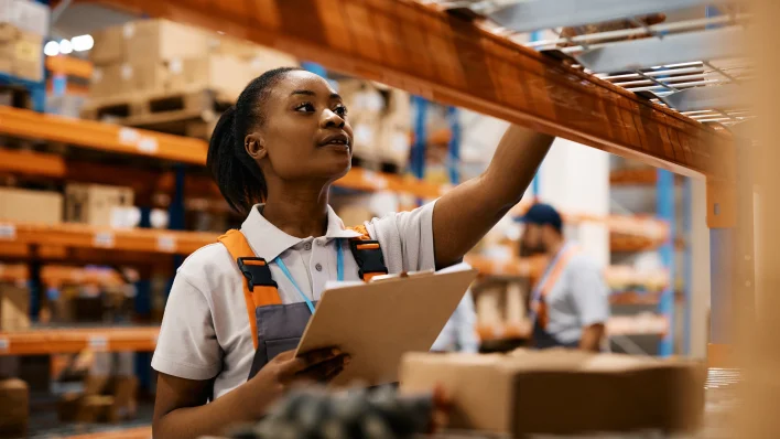 Young Black woman with clipboard reaching up to grab something from a warehouse shelf. She is wearing a shop apron and white shirt.