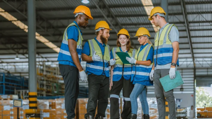 A line of 6 multicultural factory workers in hard hats and work gloves. All are pointed at the camera giving a thumbs up.