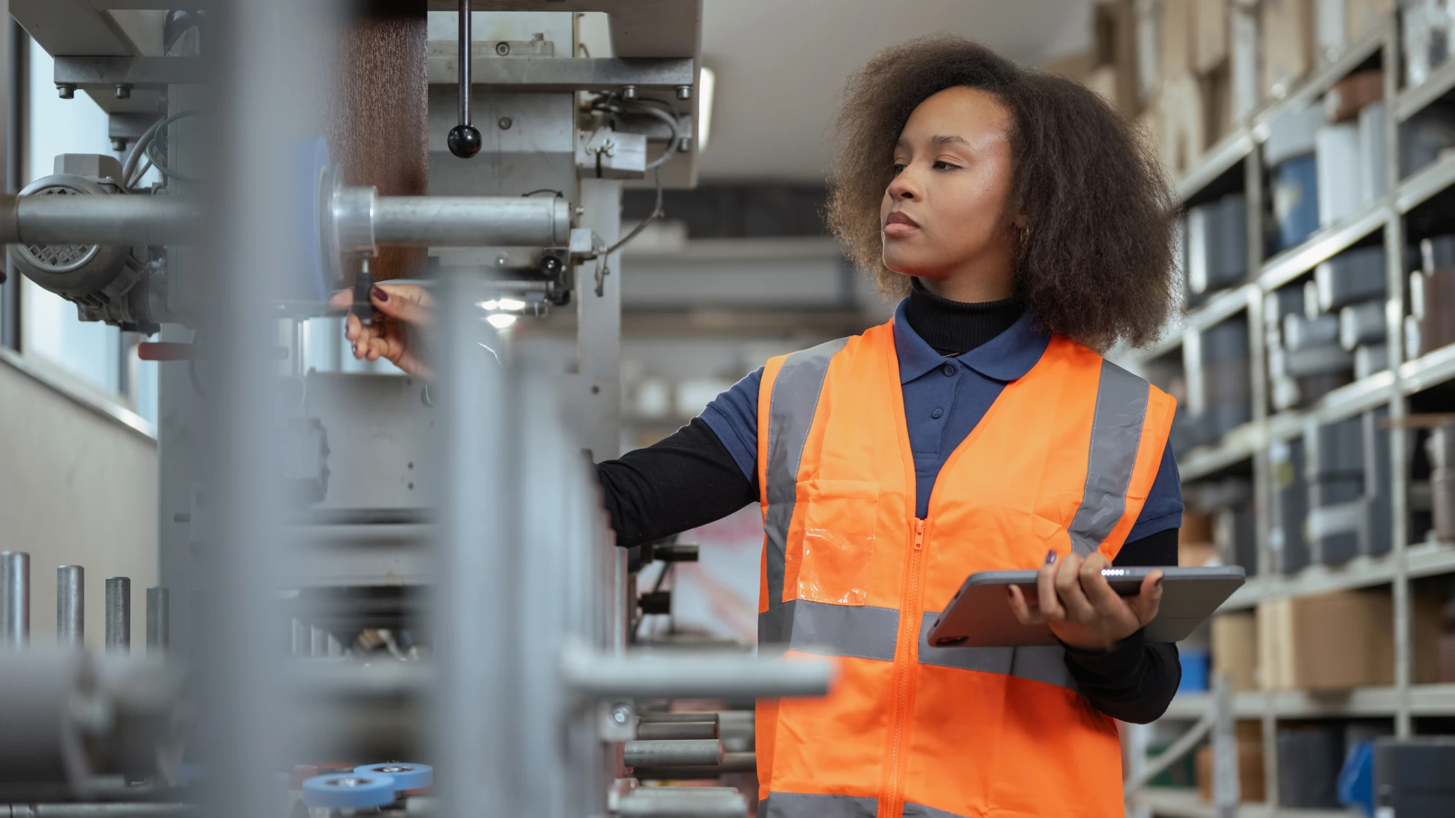 African-American woman in orange vest holds a table and adjusts a setting on a machine in an advanced manufacturing environment.
