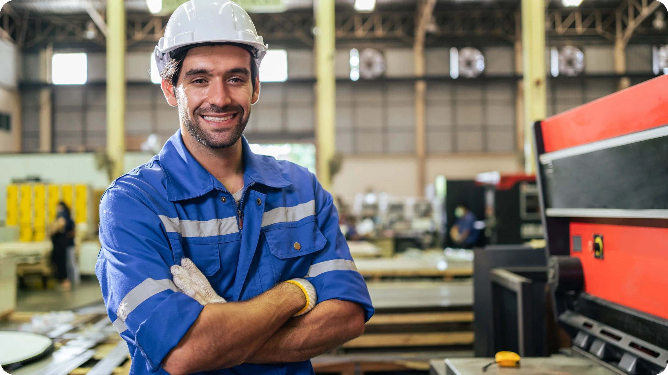 Smiling dark-haired male in hard hat and blue work coverall stands in a factory setting next to machinery.