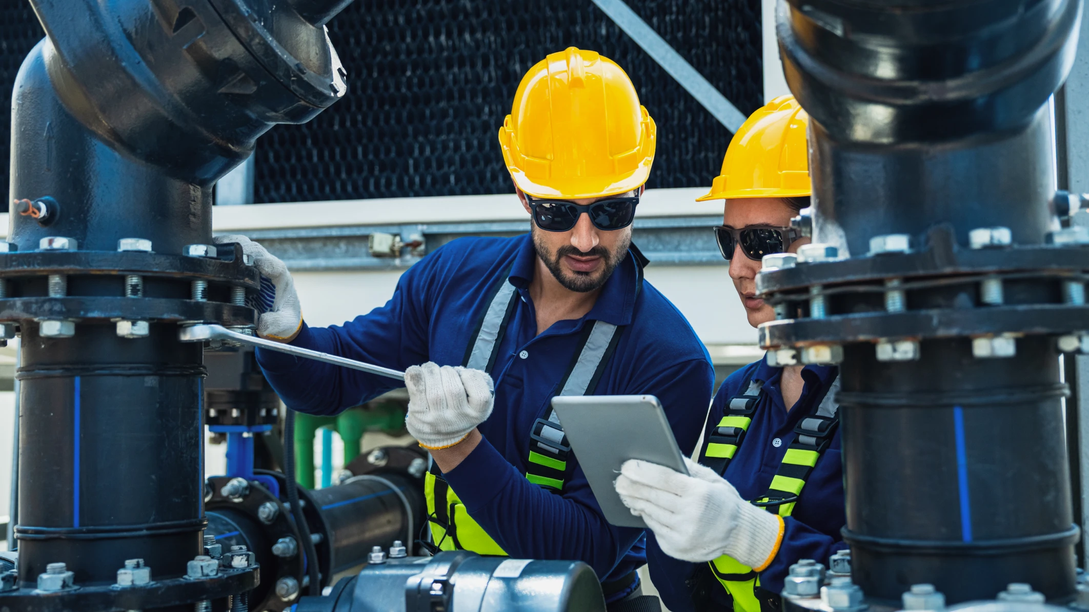 A male and female technician in yellow hard hats and safety glasses are operating automated machinery while looking at a handheld tablet.